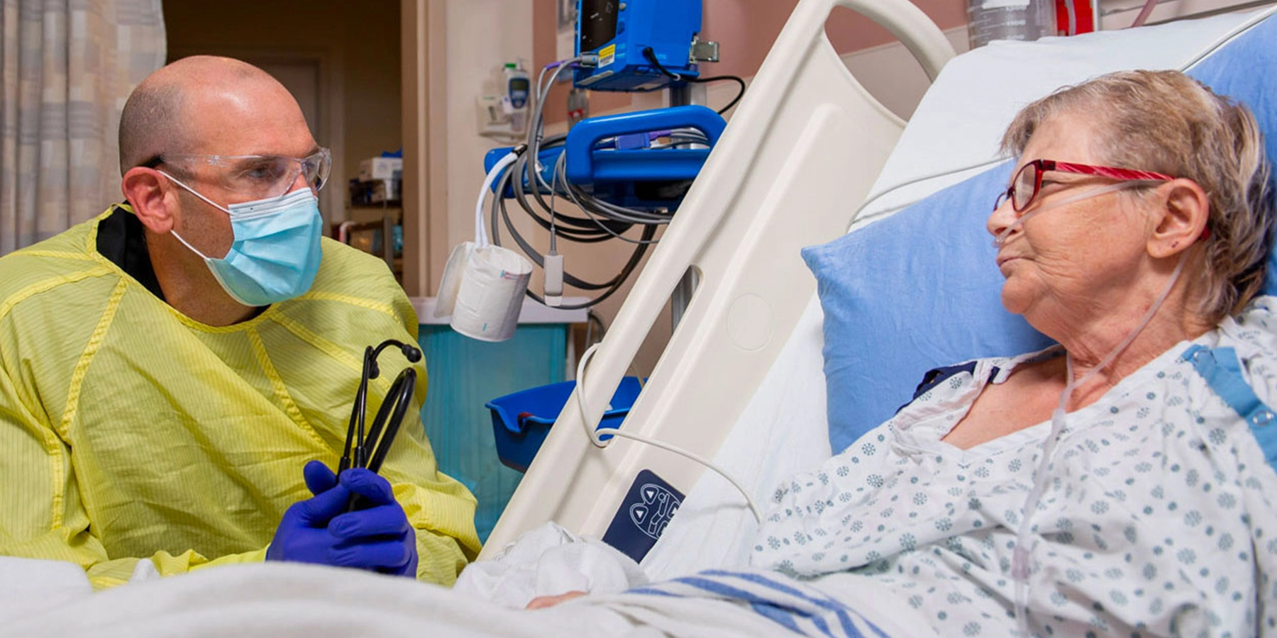 Physician in PPE sitting at the bedside of a patient.
